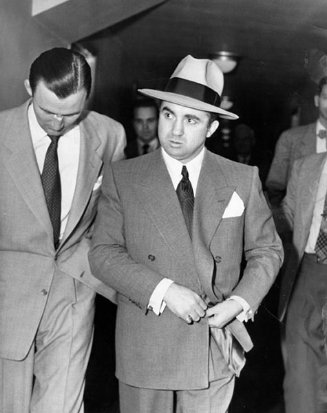 Mickey Cohen, wearing suit, tie and hat, walks indoor with a man walking closely on one side of him, and a group of men following behind.