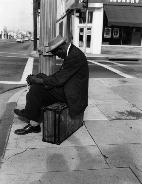 A man has fallen asleep sitting on a suitcase on a sidewalk in Los Angeles. 