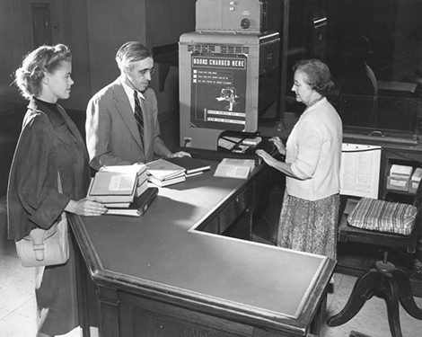 Two patrons watch as a librarian uses a Recordak machine
